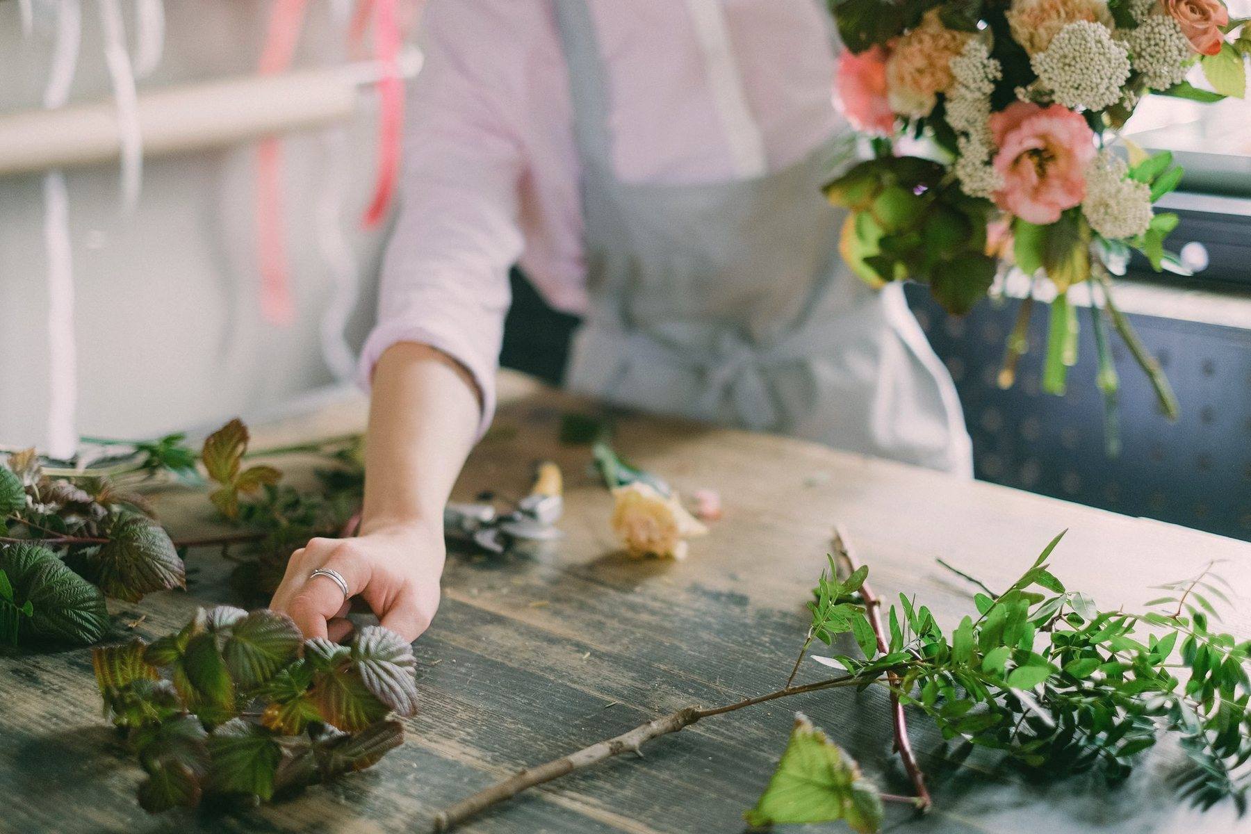 Hands arranging stems on a long wooden workshop table, a finished bouquet beside them
