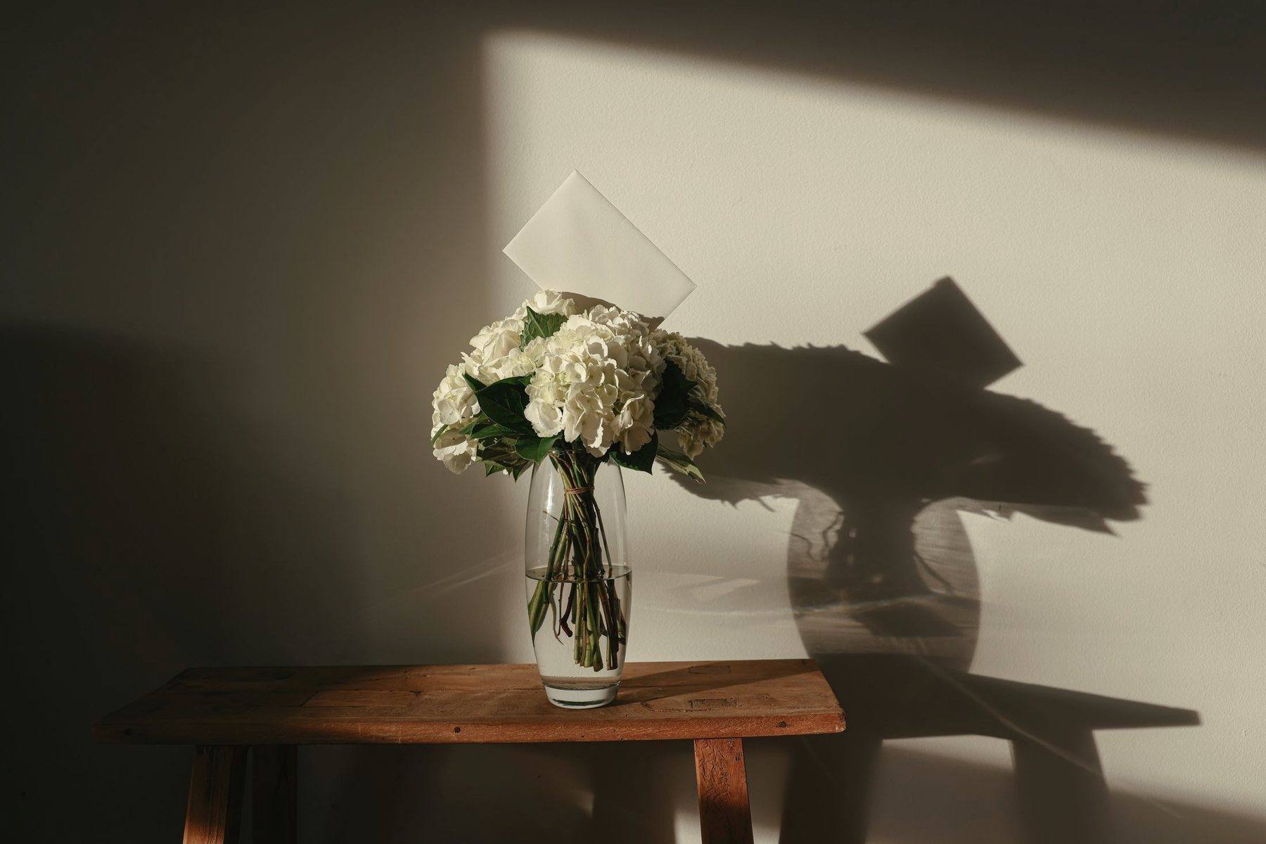 White hydrangea and roses in a glass vase on a wooden table, late sunlight casting long shadows on the wall